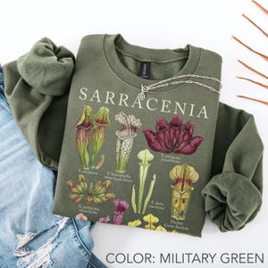 May include: A military green crewneck sweatshirt featuring a botanical illustration of Sarracenia pitcher plants. The graphic includes the text SARRACENIA and labels for various species like S. purpurea, S. leucophylla, S. alata, and S. psittacina.