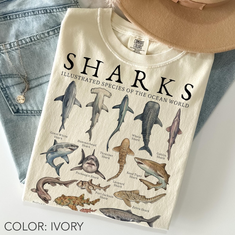 Ivory Comfort Colors unisex oversized t-shirt featuring a vintage shark species chart with illustrated great white, hammerhead, whale shark, thresher shark, goblin shark, leopard shark, sand tiger shark, zebra shark, and other ocean sharks.