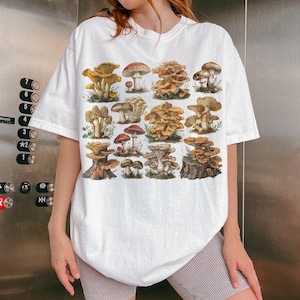 Pu&ograve; includere: T-shirt bianco con un'illustrazione dettagliata di vari funghi nei toni del marrone, rosso e giallo. Il design presenta una variet&agrave; di funghi, creando un tema botanico. La maglietta ha una vestibilit&agrave; casual e oversize.