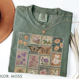 K&ouml;nnte beinhalten: Ein moosgr&uuml;nes T-Shirt mit einem Blumen- und Schmetterlingsstempel-Design. Das Shirt hat einen Rundhalsausschnitt und kurze &Auml;rmel. Eine goldene Halskette mit Anh&auml;nger liegt &uuml;ber dem Shirt. Der Text "COLOR: MOSS" steht unten.