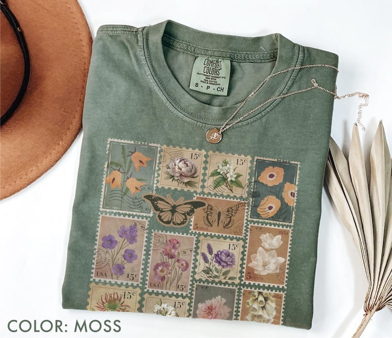 K&ouml;nnte beinhalten: Ein moosgr&uuml;nes T-Shirt mit einem Blumen- und Schmetterlingsstempel-Design. Das Shirt hat einen Rundhalsausschnitt und kurze &Auml;rmel. Eine goldene Halskette mit Anh&auml;nger liegt &uuml;ber dem Shirt. Der Text "COLOR: MOSS" steht unten.