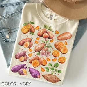 May include: Cream-coloured t-shirt with a watercolour design of sweet potatoes, including orange and purple varieties, with green leaves and orange slices. The shirt has a crew neck and the text "COLOR: IVORY" at the bottom.