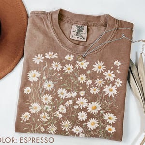 May include: Espresso-colored t-shirt with a daisy floral design. The shirt has a crew neck and short sleeves. A silver necklace with a pendant is draped across the shirt. The shirt is made of a soft material.