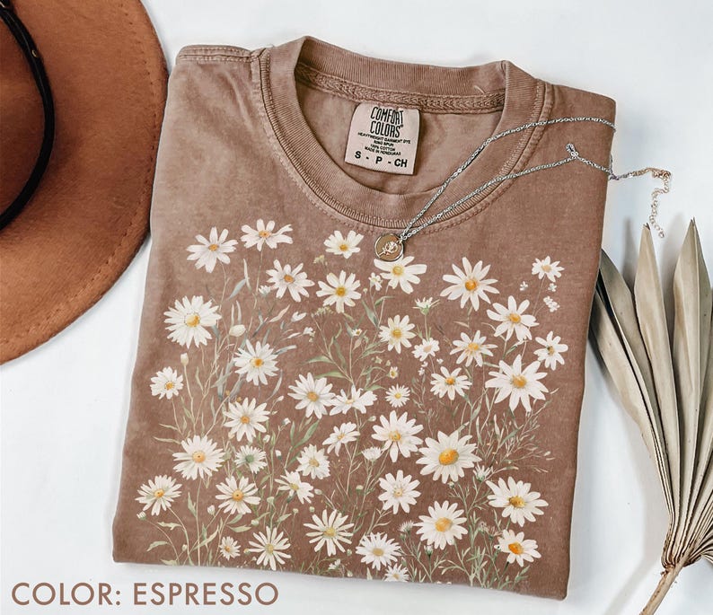 May include: Espresso-colored t-shirt with a daisy floral design. The shirt has a crew neck and short sleeves. A silver necklace with a pendant is draped across the shirt. The shirt is made of a soft material.