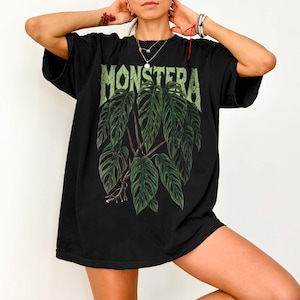 May include: A black short-sleeved t-shirt featuring a large graphic print of a green Monstera plant with detailed leaves and roots. The word MONSTERA is printed in a distressed, light green font above the plant illustration on the front chest.