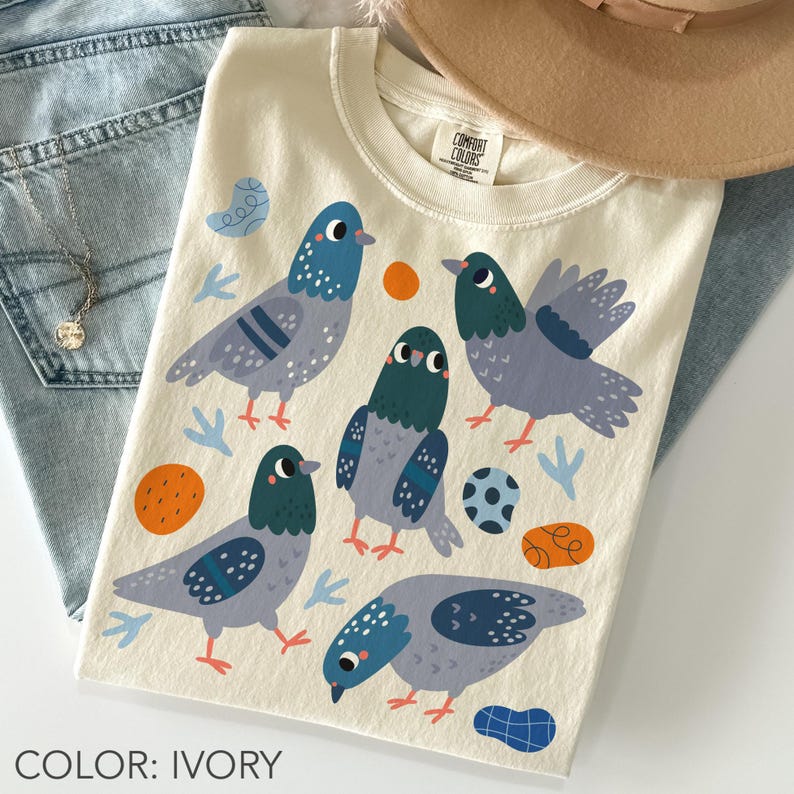 May include: Ivory-colored t-shirt featuring a playful design of several cartoon pigeons in shades of blue, gray, and teal. The birds are surrounded by orange and blue abstract shapes. The text "COLOR: IVORY" is at the bottom.