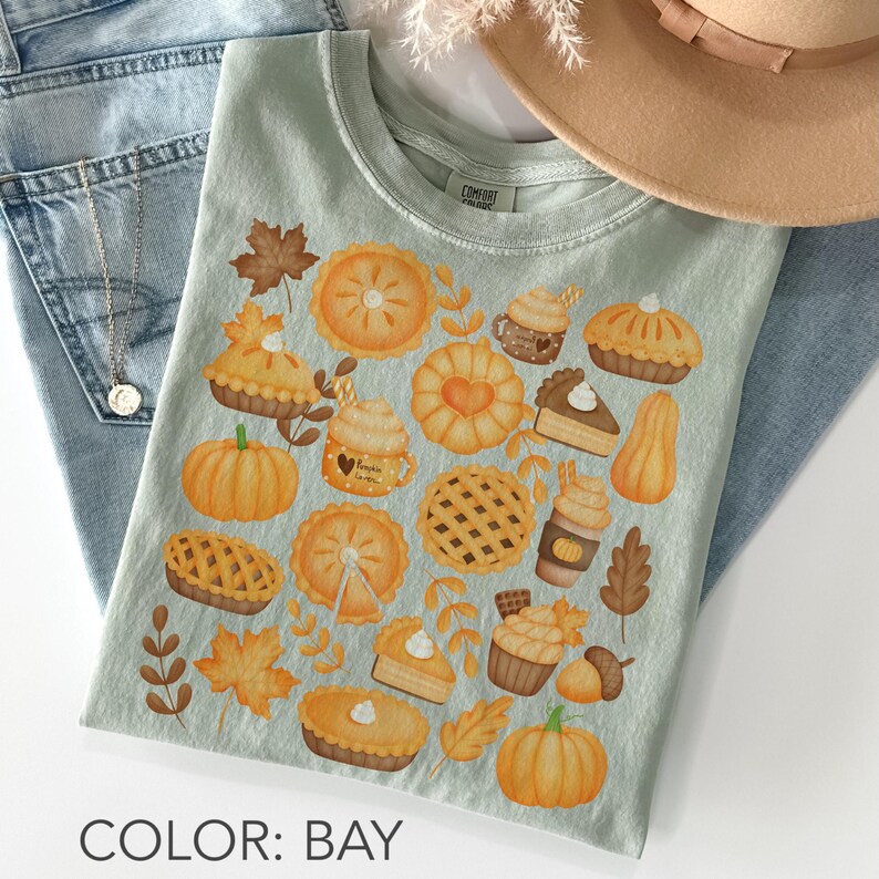 May include: A pale green t-shirt with a fall design featuring pumpkins, pies, and autumn leaves. The shirt has a crew neck and short sleeves. The text "COLOR: BAY" is at the bottom of the image.