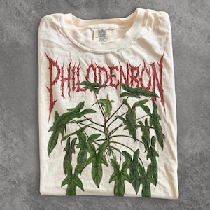 May include: A cream-colored t-shirt featuring a graphic of a green plant with lobed leaves. Above the plant, the word PHILODENDRON is printed in a distressed, jagged red font. The shirt has a crew neckline and a Comfort Colors brand label.