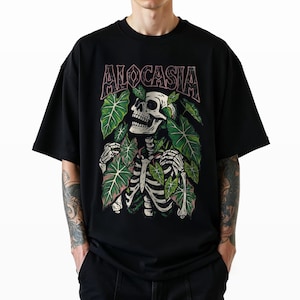 K&ouml;nnte beinhalten: Schwarzes T-Shirt mit einem Skelett-Design, umgeben von gr&uuml;nen Bl&auml;ttern. Das Wort "ALOCASIA" ist in einer stilisierten Schriftart &uuml;ber dem Skelett aufgedruckt. Das Design hat ein botanisches und gotisches Thema.