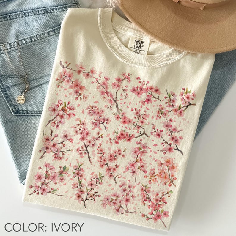 May include: Ivory-colored t-shirt featuring a watercolor design of pink cherry blossom branches and flowers. The shirt is a crew neck style and is paired with blue jeans and a necklace. The text "COLOR: IVORY" is at the bottom.