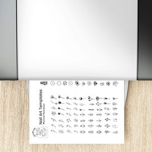 A printable nail art template sheet coming out of a printer, showing rows of hand-drawn floral and botanical nail icons in black line art. The design sheet includes flowers, petals, leaves, branches, and tiny botanical shapes arranged neatly for easy tracing or stenciling. This image illustrates how customers can instantly download, print, and use the nail art templates to create DIY floral nail designs for gel nails or press-on nails.