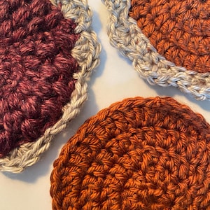 May include: Close-up of three handmade crochet coasters. One is burgundy with a gray scalloped edge, and two are shades of orange with gray edges. The coasters are round and have a textured appearance, perfect for protecting surfaces.
