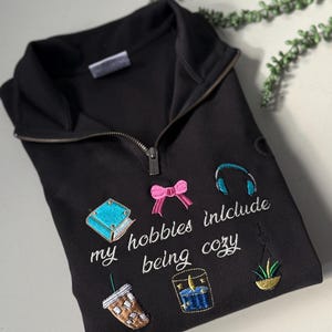 Puede incluir: Sudadera negra con cremallera de cuarto de cierre con imágenes bordadas de un libro, un lazo, auriculares, café, una vela y una planta colgante. El texto "my hobbies include being cozy" está bordado en blanco.