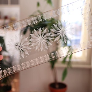 Könnte beinhalten: Ein klares, rechteckiges Serviertablett aus Glas mit einem dekorativen weißen Blumen- und geometrischen Muster. Das Tablett hat drei große Blumendesigns und einen Rand aus sich wiederholenden Formen. Das Tablett wird vor einem Fenster gehalten.