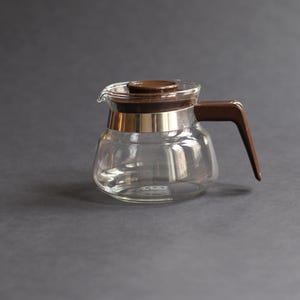 Puede incluir: Una cafetera de vidrio transparente con asa y tapa marrones. La cafetera tiene una banda dorada alrededor del centro. El asa es angular y la tapa es redonda. La cafetera está vacía y se encuentra sobre una superficie gris.