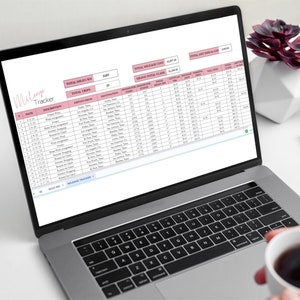 May include: A laptop computer screen displaying a mileage tracker spreadsheet with columns for date, description, miles, and total miles. The spreadsheet is titled "Mileage Tracker" and has a pink background. The spreadsheet is on a white surface with a cup of coffee and a pair of glasses.