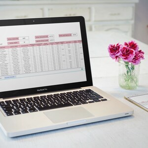 May include: A silver laptop computer with a spreadsheet open on the screen. The spreadsheet is titled "Mileage Tracker" and contains columns for date, destination, total miles driven, and other information. The laptop is sitting on a white table with a vase of pink flowers and a white smartphone.