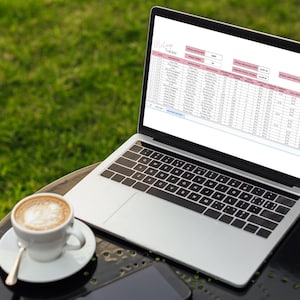 May include: A laptop computer with a spreadsheet open on the screen. The spreadsheet is titled "Affiliate Tracker" and contains data about social media marketing. The laptop is on a black table with a cup of coffee and a smartphone.