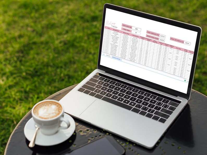 May include: A laptop computer with a spreadsheet open on the screen. The spreadsheet is titled "Affiliate Tracker" and contains data about social media marketing. The laptop is on a black table with a cup of coffee and a smartphone.