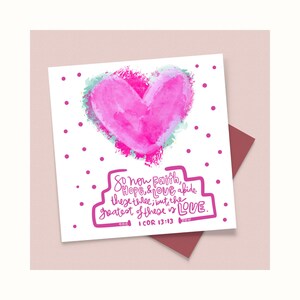 Könnte beinhalten: Eine Grußkarte mit einem rosa Aquarellherz und dem Text "So now Faith, Hope, & LOVE abide these three; but the greatest of these is LOVE." Die Karte liegt auf einem weinroten Umschlag mit rosa Punkten.