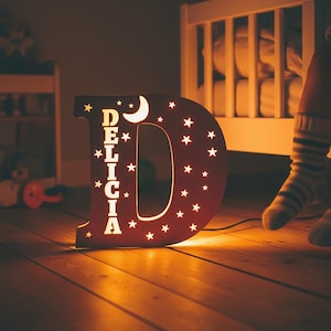 Op de afbeelding: Een mahoniehouten nachtlampje in de vorm van de letter "D" met de naam "DELICIA" uitgesneden. De letter is versierd met sterren en een halve maan. Het licht is aan en verlicht de kamer. Het licht staat op een houten vloer.