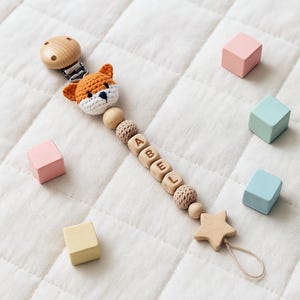 Puede incluir: Un clip de chupete de bebé de madera con el nombre "ABEL" escrito con cuentas de letras de madera. El clip presenta una cabeza de zorro de ganchillo en naranja y blanco, cuentas de madera y un mordedor en forma de estrella. Bloques de madera de colores pastel están dispersos.