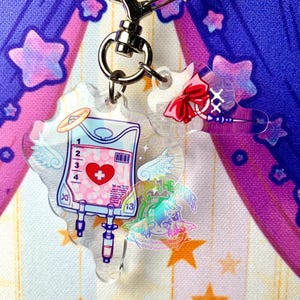 May include: A clear acrylic keychain featuring a pink and blue IV bag with a red heart and a white cross. The IV bag has a white label with the text "50 ml". The keychain also includes a pink bow with a white cross and a white angel wing on each side of the IV bag.