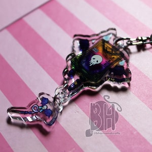 May include: A clear acrylic charm featuring a rainbow-coloured book with a skull and bats, and a wand with a purple dragon. The charm is attached to a silver chain.