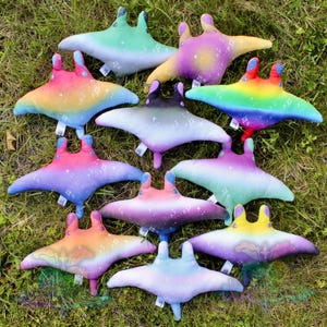 May include: A collection of ten plush manta ray toys featuring various gradient color patterns, including rainbow, sunset, and galaxy themes. Each soft toy displays small white star and sparkle graphics across the fabric surface.