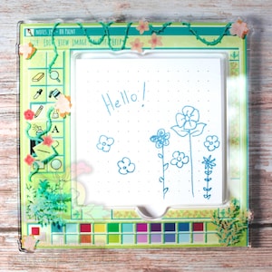 Puede incluir: Un marco acrílico transparente con una inserción de papel cuadriculado blanco. El papel tiene un dibujo floral azul y la palabra "Hello!". El marco tiene un borde verde con detalles florales y de vid, y un diseño de paleta de pintura colorida.