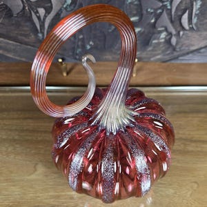 May include: A decorative glass pumpkin with a swirling stem. The pumpkin is a deep red color with white speckles and a clear, curved stem. The glass has a ribbed texture.