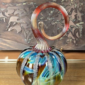 May include: A glass pumpkin with a swirling, multi-colored design in shades of blue, brown, and gold. The stem is a spiral of red and gold glass.