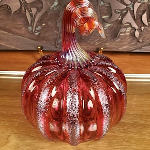 May include: A red glass pumpkin with a swirling stem. The pumpkin has a speckled, iridescent finish.