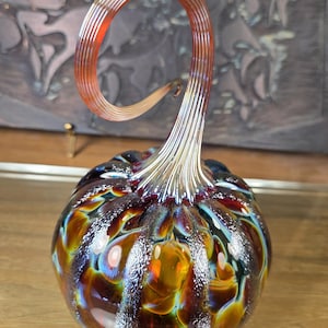 May include: A hand-blown glass pumpkin sculpture featuring a rounded body with swirling patterns of amber, deep brown, and iridescent blue hues. The piece is topped with a tall, ribbed, translucent orange stem that curves gracefully at the top.