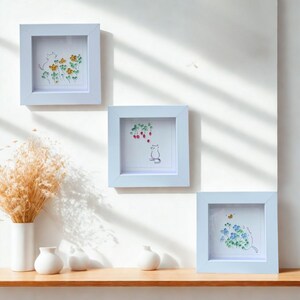 Puede incluir: Tres obras de arte enmarcadas con marcos cuadrados de color azul claro. Cada marco contiene una ilustración caprichosa de un gato con flores, bayas o una abeja. Las obras de arte se exhiben en una pared blanca sobre una estantería de madera con jarrones blancos.