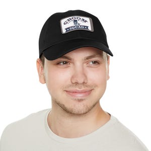 Puede incluir: Gorra de béisbol negra con un parche rectangular que dice "GROOM SQUAD" en blanco y azul marino. La gorra está hecha de un material suave y tiene una visera curva. El parche presenta un gráfico de una silla de barbero.