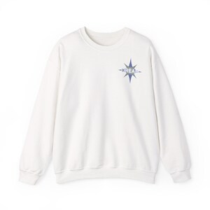 Puede incluir: Una sudadera blanca de cuello redondo con mangas largas. Un gráfico de estrella azul con la palabra "DREAM" está impreso en la parte superior izquierda del pecho. La sudadera está hecha de un material suave.