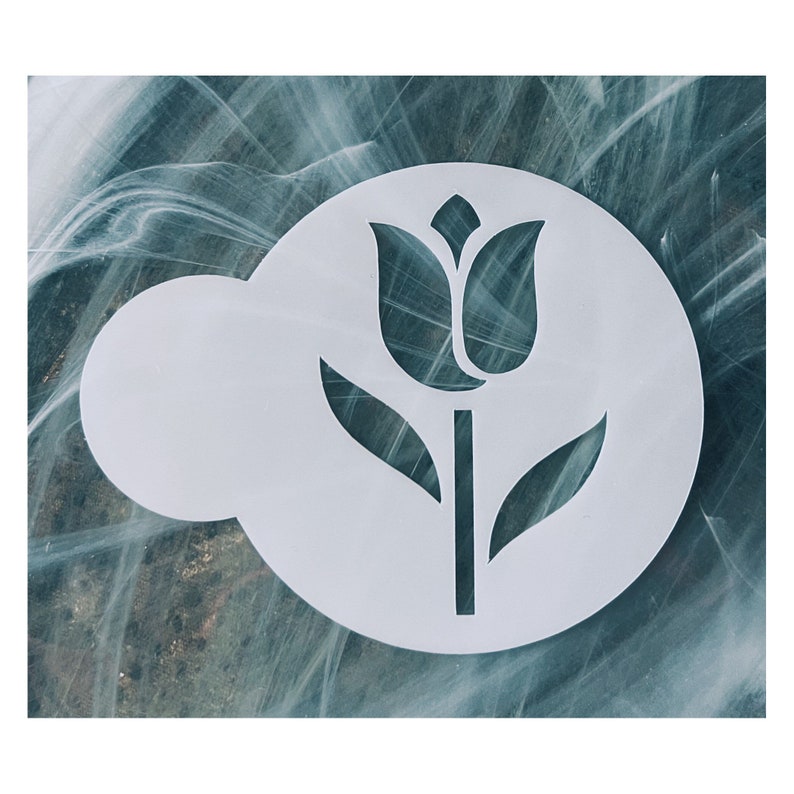 May include: A white, circular stencil featuring a tulip design. The stencil has a flower with two leaves and a stem. The background is a marbled pattern of teal, white, and gray.