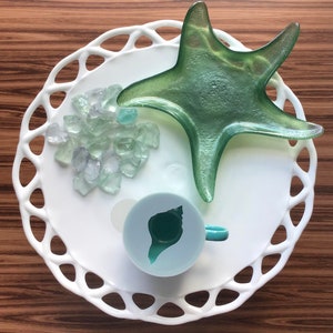May include: A white ceramic cake stand with a scalloped edge, featuring a green glass starfish, a pile of clear glass sea glass, and a teal blue seashell-shaped mold.