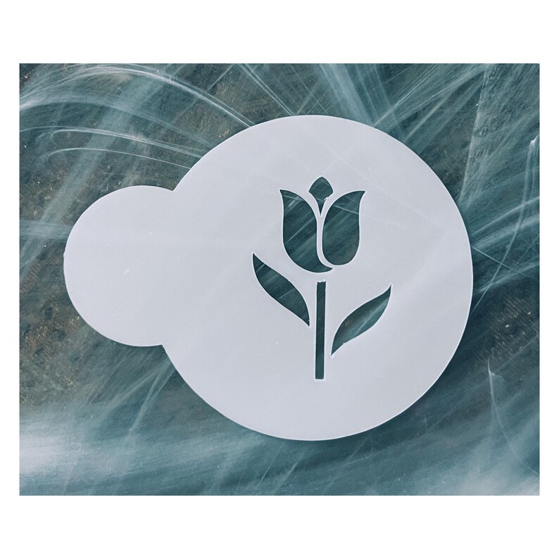May include: A white stencil featuring a tulip design. The stencil is circular with a cloud-shaped extension on the left side. The tulip has a detailed flower head, stem, and leaves. The stencil is designed for crafting and decorative purposes.