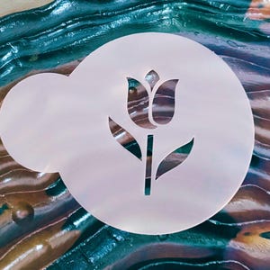May include: A white, circular stencil featuring a tulip design. The stencil has a rounded shape with a smaller, attached section. The tulip design includes a stem, leaves, and the flower's petals. The background has a colorful, abstract pattern.
