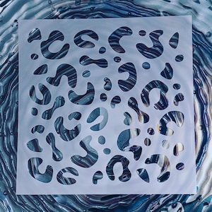 May include: A square, semi-transparent stencil featuring a repeating leopard print pattern with various organic, curved shapes and small circular cutouts, layered over a background of swirling blue, black, and metallic silver paint.