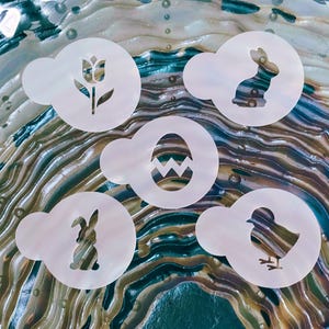 May include: Five white plastic stencils with Easter-themed designs. The stencils feature a tulip, a bunny, an Easter egg, another bunny, and a chick. The stencils are circular with a small handle and are set against a colorful, swirled background.