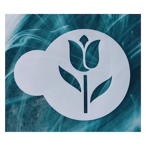 May include: A white stencil featuring a tulip design. The stencil is circular with a cloud-shaped extension on the left side. The tulip has a stem, leaves, and a detailed flower head. The background is a teal and white marbled pattern.