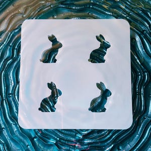May include: A white silicone mold with four bunny-shaped cavities. The bunnies are dark green with a marbled effect. The background is a textured, swirling pattern in shades of blue and green.