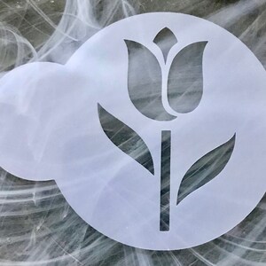 May include: A white, circular stencil featuring a tulip design. The tulip is depicted with a stem, leaves, and a detailed flower head. The stencil is designed for crafting and decorating projects.