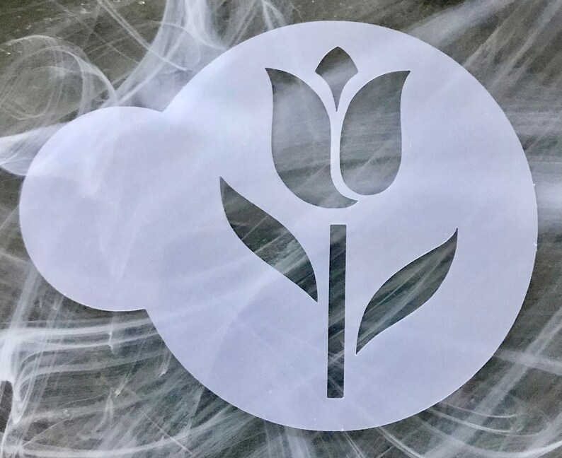 May include: A white, circular stencil featuring a tulip design. The tulip is depicted with a stem, leaves, and a detailed flower head. The stencil is designed for crafting and decorating projects.