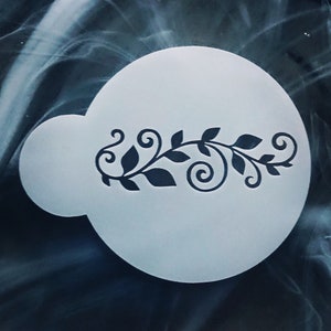 May include: White plastic stencil with a black floral design. The design features a vine with leaves and swirls.