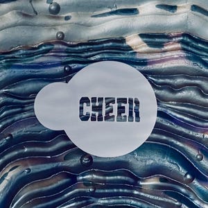 May include: A circular white stencil featuring the word CHEER in a bold, blocky font. The stencil has a small circular tab on the left side and is positioned over a dark, wavy, iridescent surface with blue, purple, and metallic gray tones.