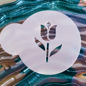 May include: A white, circular stencil featuring a tulip design. The stencil has a rounded shape with a smaller, attached section. The tulip design is cut out, revealing the background. The background has a mix of blue, green, and brown colors.